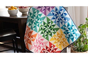 Spinning Swallows Table Topper quilt in rainbow colors draped over a chair, featuring the Flying Swallows block made with the GO!® Flying Swallows Die.