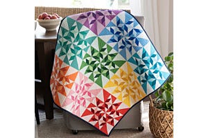 Spinning Swallows Table Topper quilt in rainbow colors draped over a chair, featuring the Flying Swallows block made with the GO!® Flying Swallows Die.