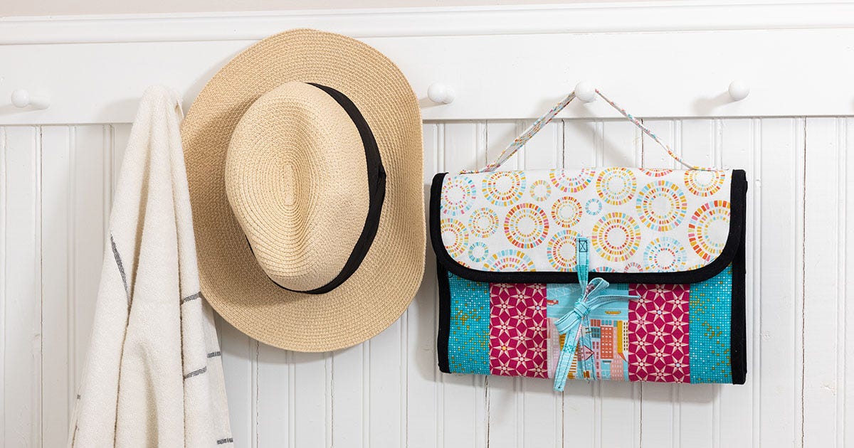 Colorful, pink and blue quilt-as-you-go travel organizer bag displayed against a white wood background on a hook next to a summer straw hat and beach towel. 