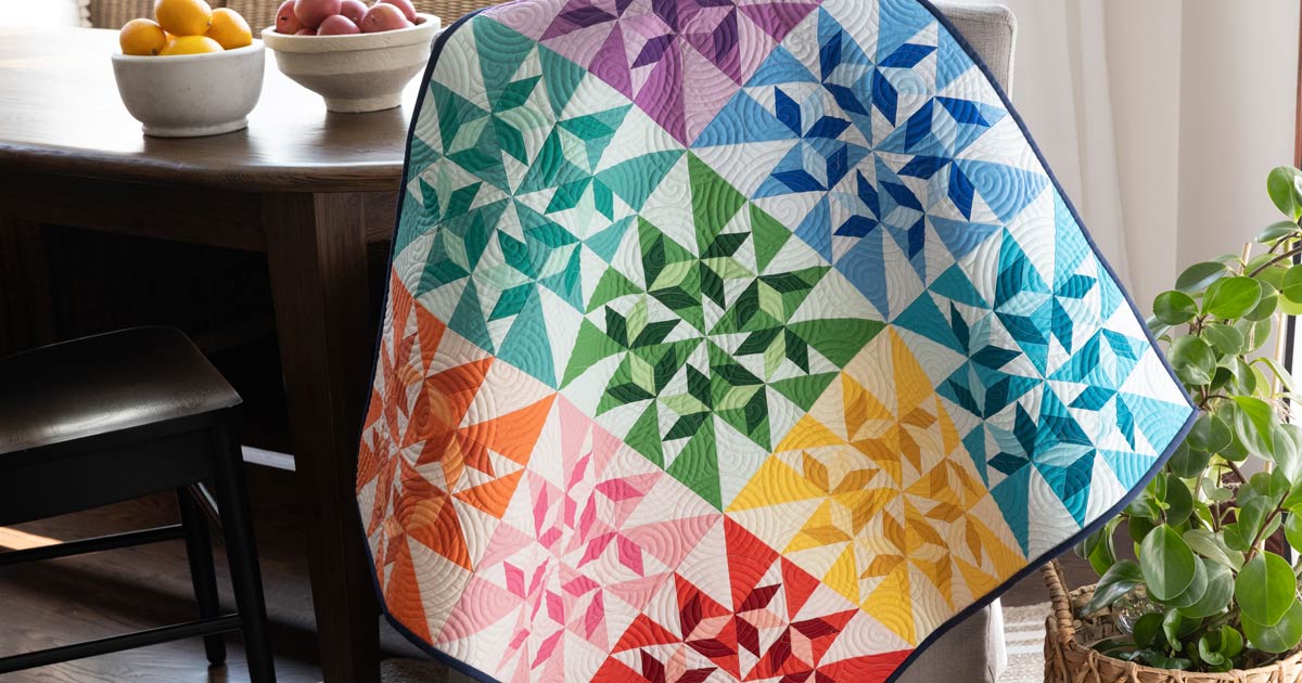 Spinning Swallows Table Topper quilt in rainbow colors draped over a chair, featuring the Flying Swallows block made with the GO!® Flying Swallows Die.