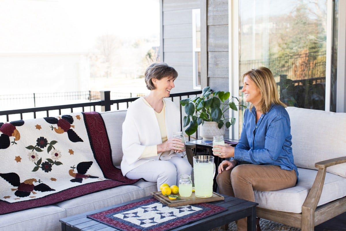 Two women sit outside next to a properly cared for quilt. They are enjoying cups of lemonade over a friendly discussion.